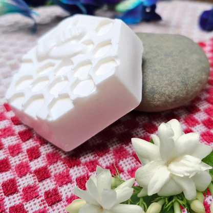 A bar of Mogra soap placed on a red and white checkered surface, with a rock and a jasmine flower in the background.